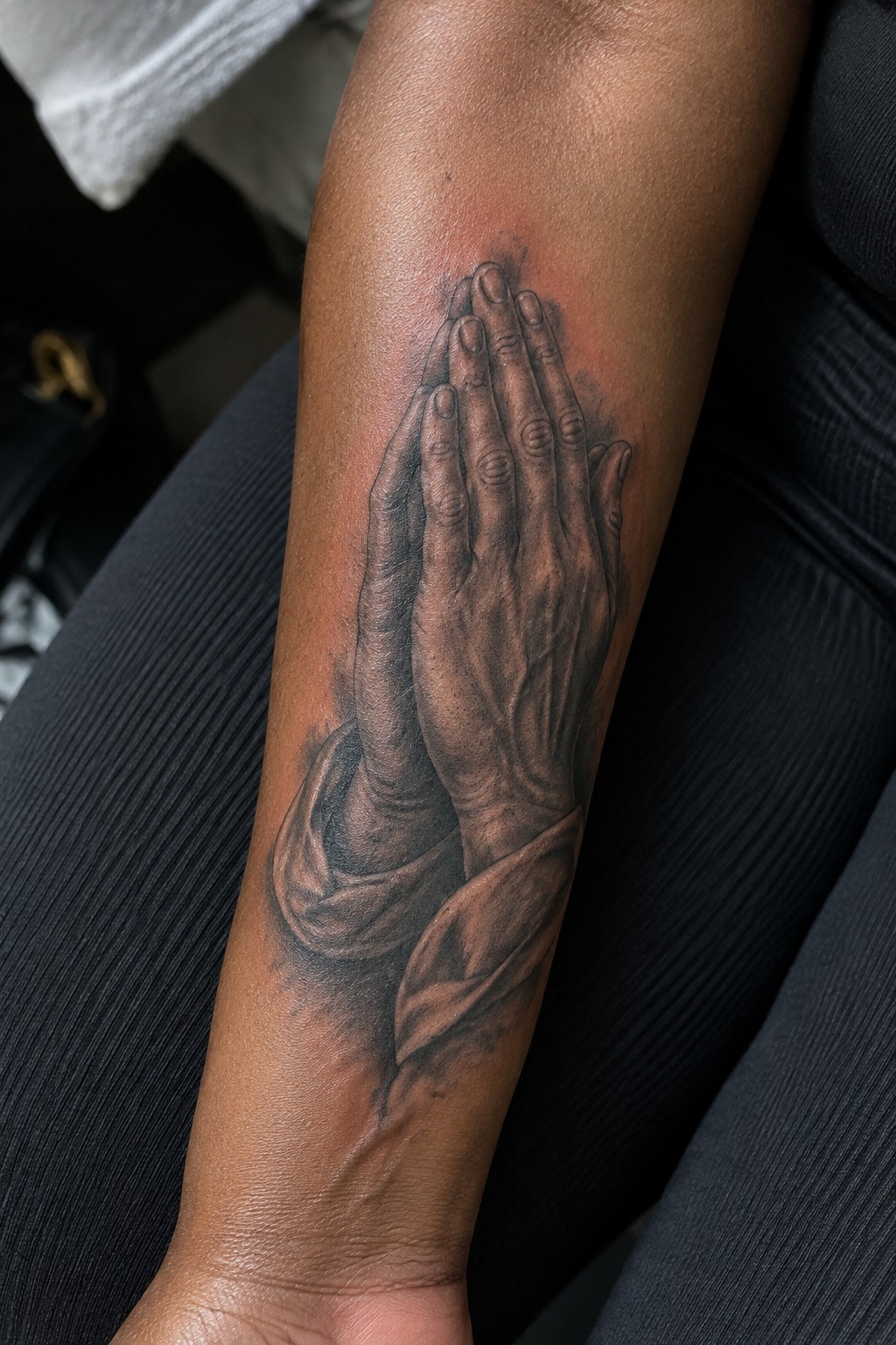 Realistic black and gray praying hands tattoo on dark-skinned forearm with red shading.