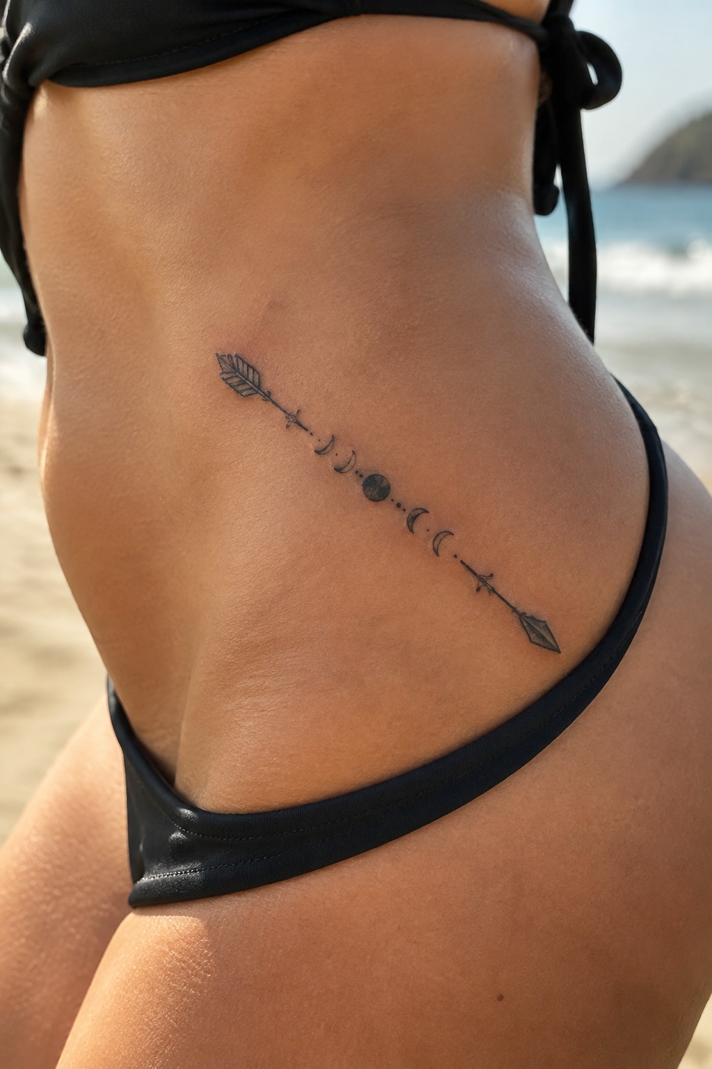 Fine-line arrow tattoo with moon phases across a woman’s hip at the beach.