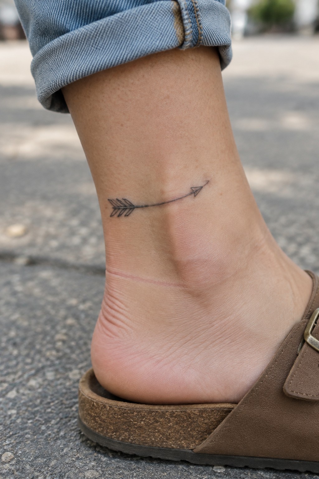 Small black fine-line arrow tattoo curving across the outer ankle above a brown sandal.