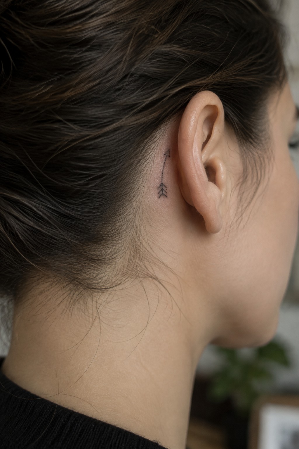 Small fine-line arrow tattoo behind a woman’s ear, with a dotted shaft and simple feather details.