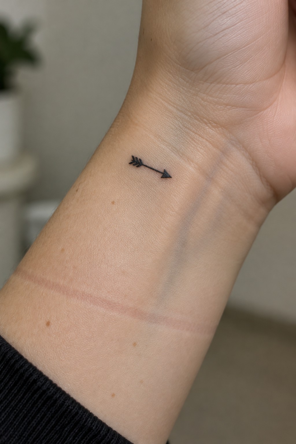 Small black arrow tattoo on the inner wrist, with a feathered tail and arrowhead.