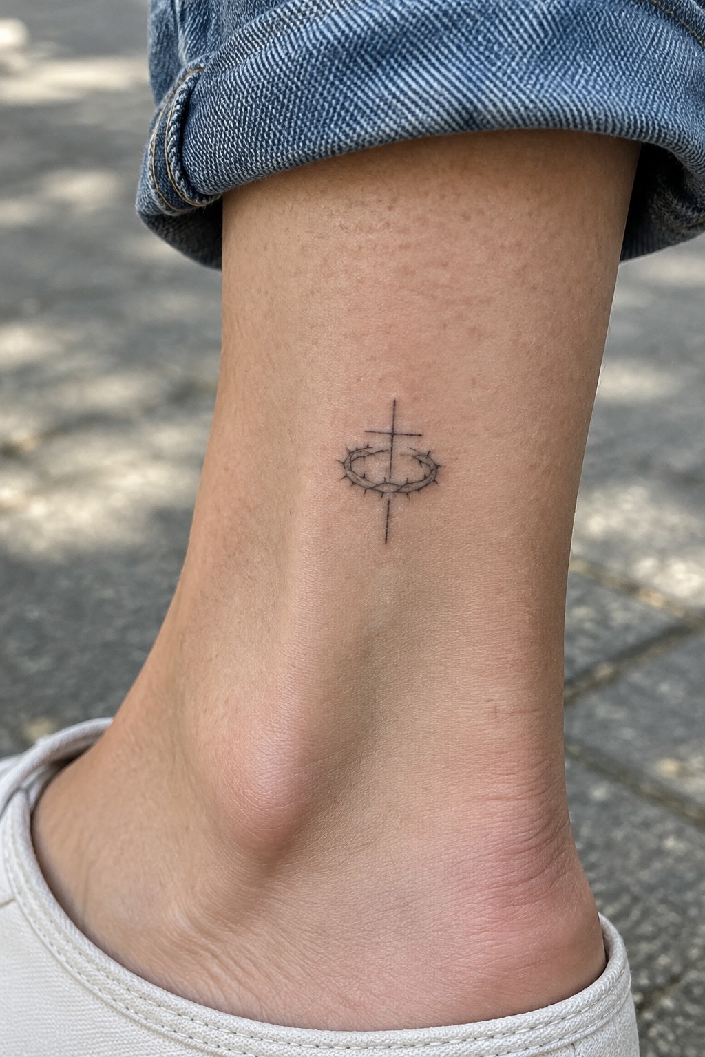 Black cross with crown of thorns tattoo on ankle below rolled blue jeans