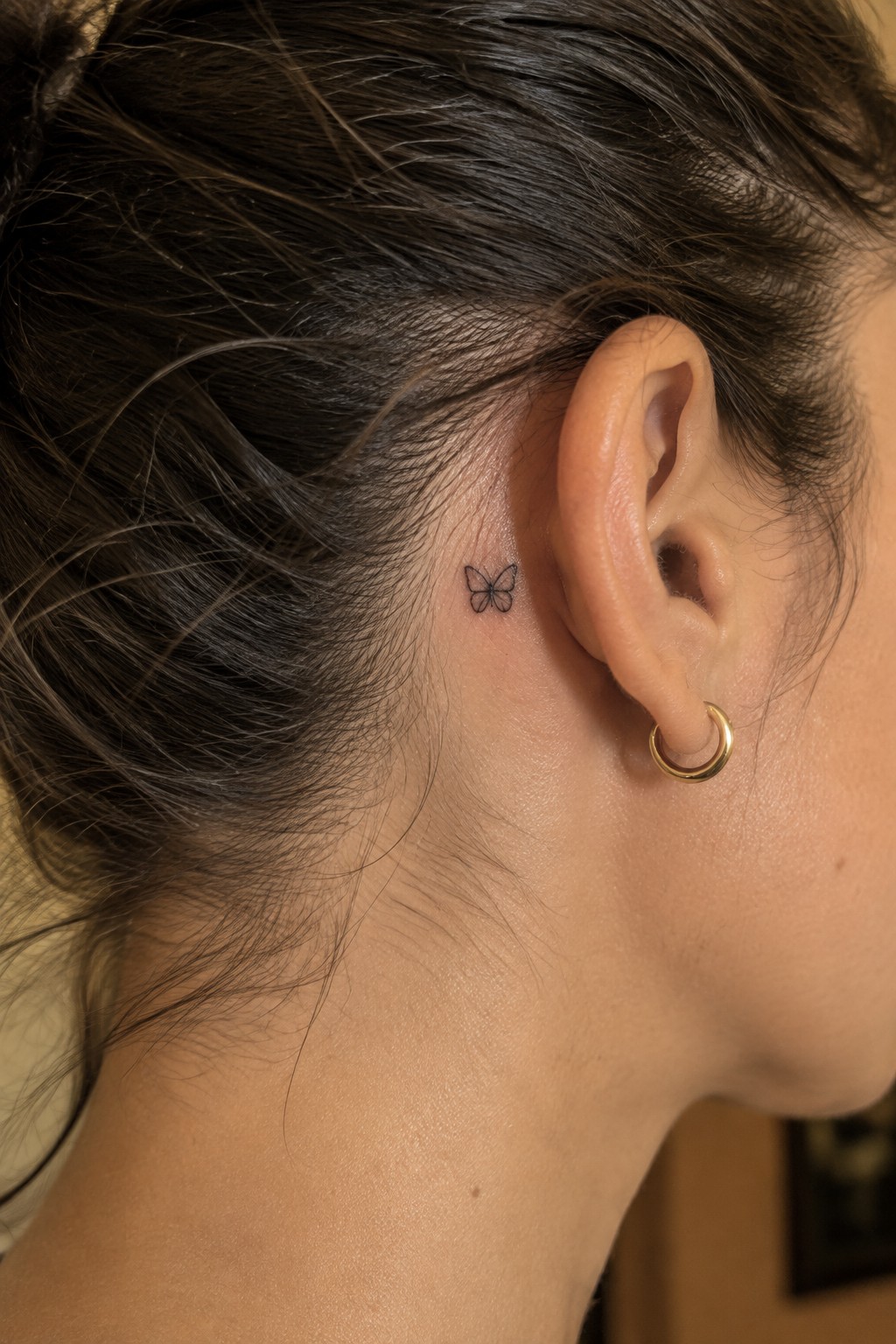 Small fine-line butterfly tattoo behind a woman’s ear, above a gold hoop earring.