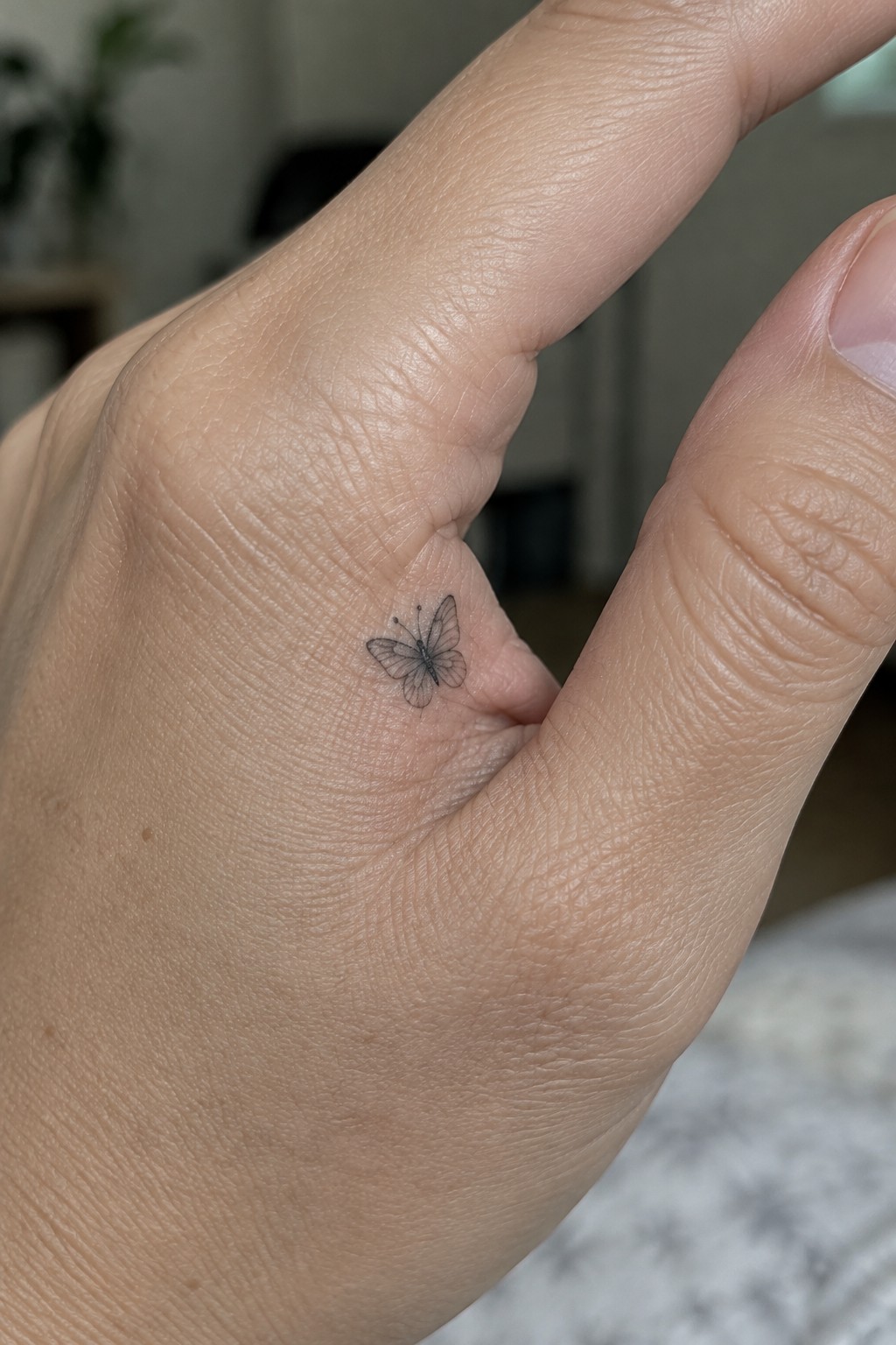 Small fine-line butterfly tattoo in the web of the hand near the thumb.
