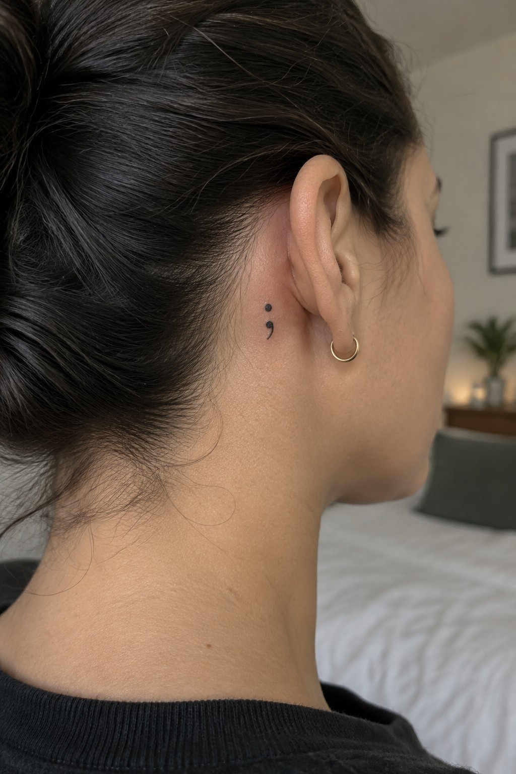Small black semicolon tattoo behind a woman’s ear, above a gold hoop earring.