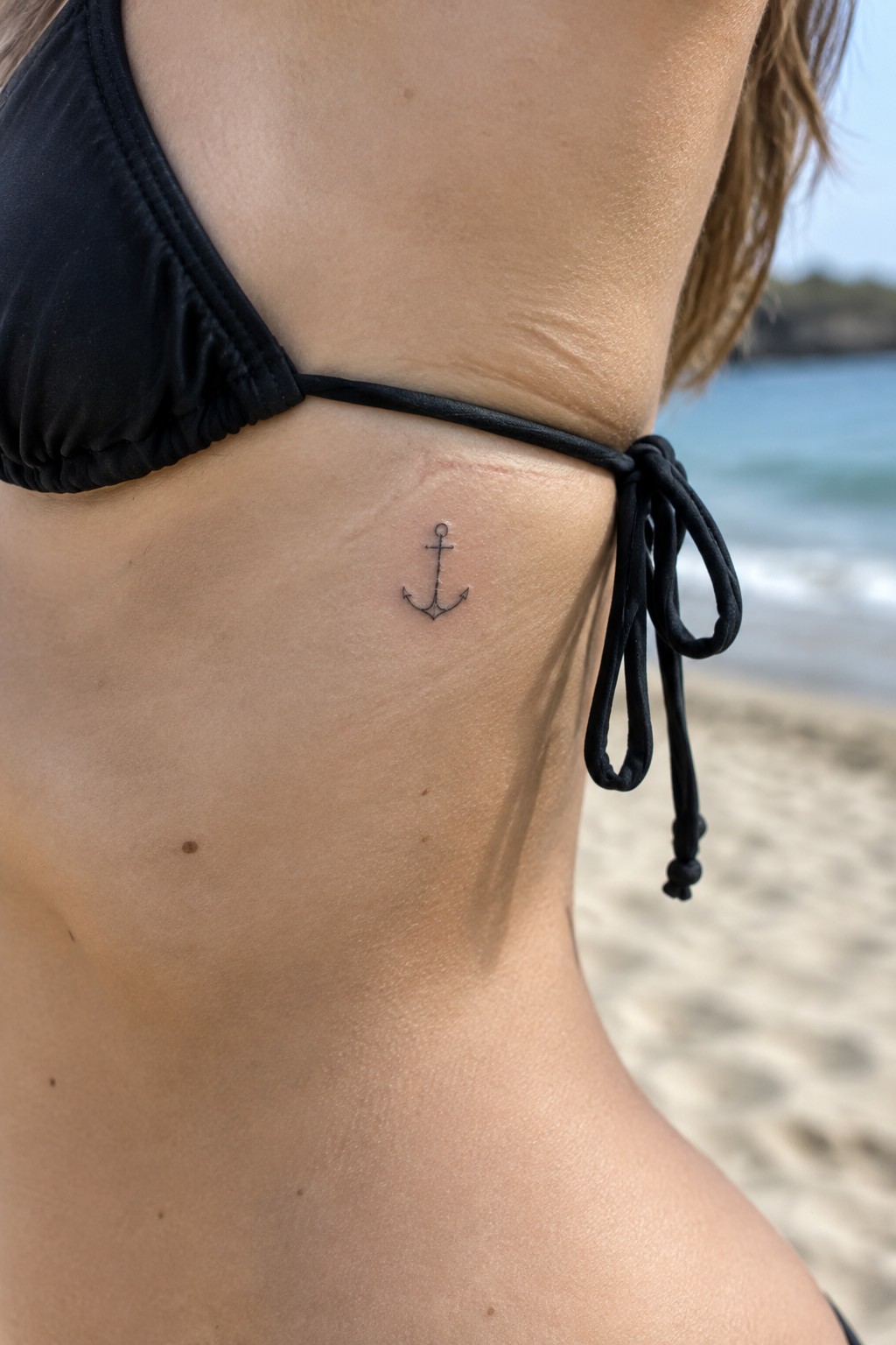 Small fine-line black anchor tattoo on the side of a woman’s torso, just below a black bikini top.