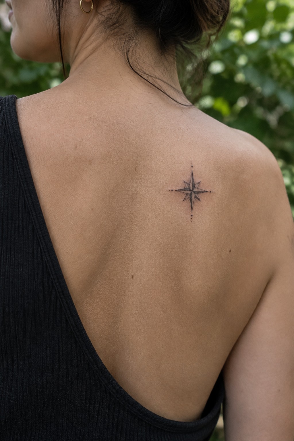 Small shaded compass rose tattoo on a woman’s upper back near the shoulder.