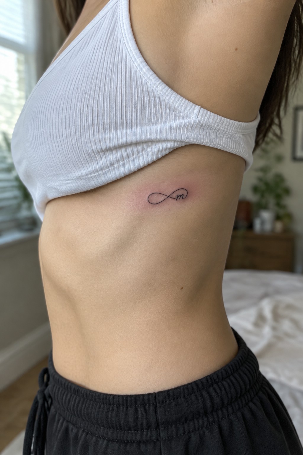 Small black infinity tattoo with a cursive initial on a woman’s side below a white top.
