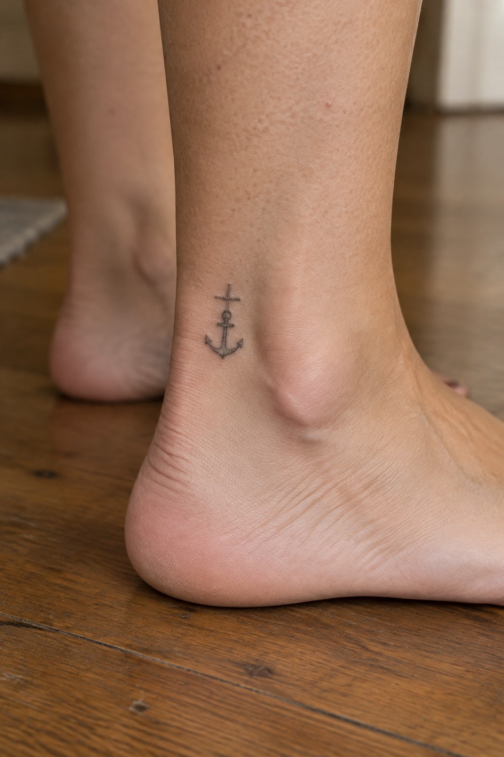 Small fine line anchor tattoo with a cross on top, placed on the inner ankle of a woman's foot.