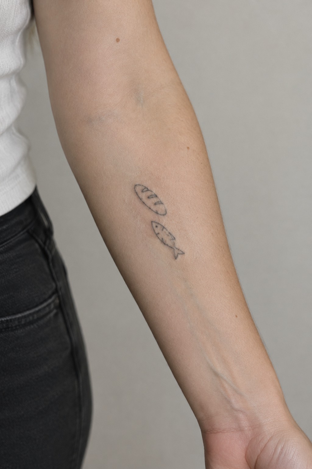 Fine line tattoo of two fish stacked vertically on the inner forearm, one rounded shape above a smaller fish with visible fins.