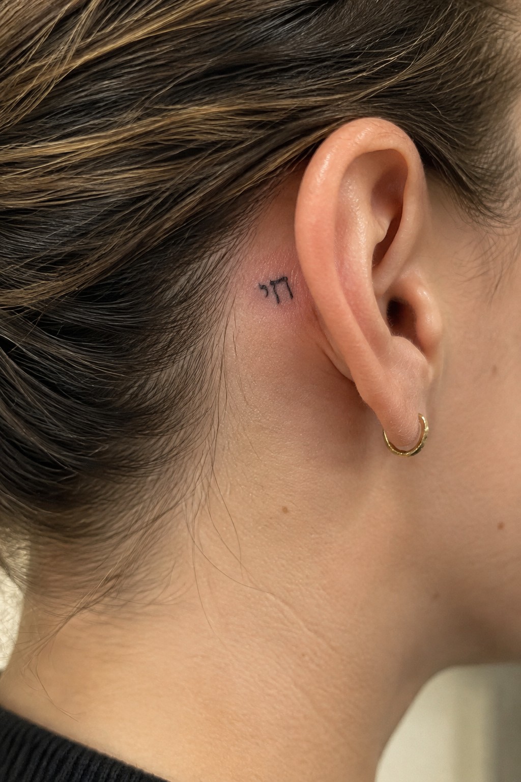 Small Hebrew tattoo behind ear beside gold hoop earring on brown hair