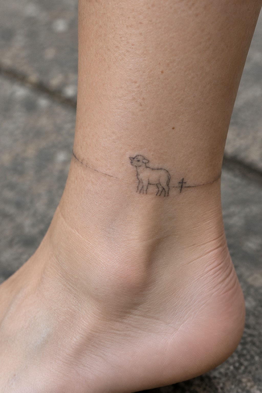 Fine line tattoo of a lamb with a thin line and small cross placed horizontally on the ankle.
