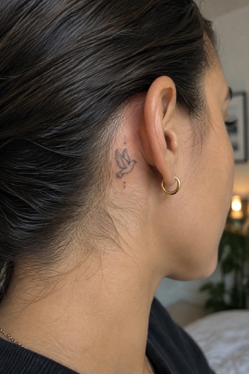 Fine line minimalist dove tattoo placed behind the ear on dark hair, shown from the side profile