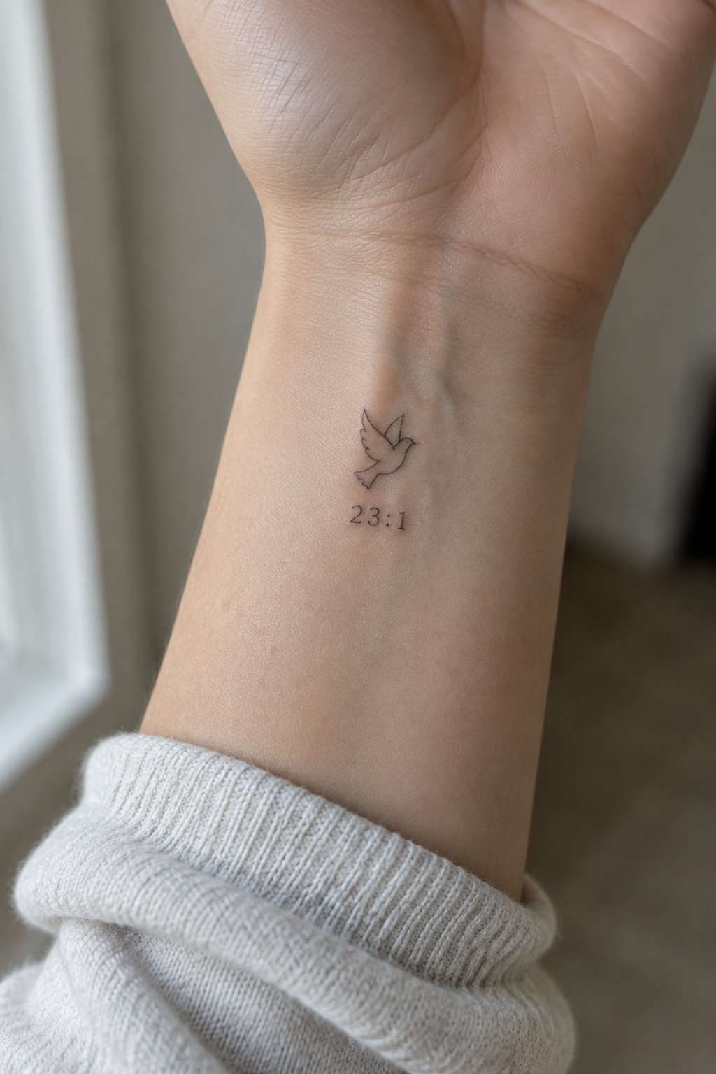 Tiny bird tattoo with 23:1 on inner wrist above white knit cuff