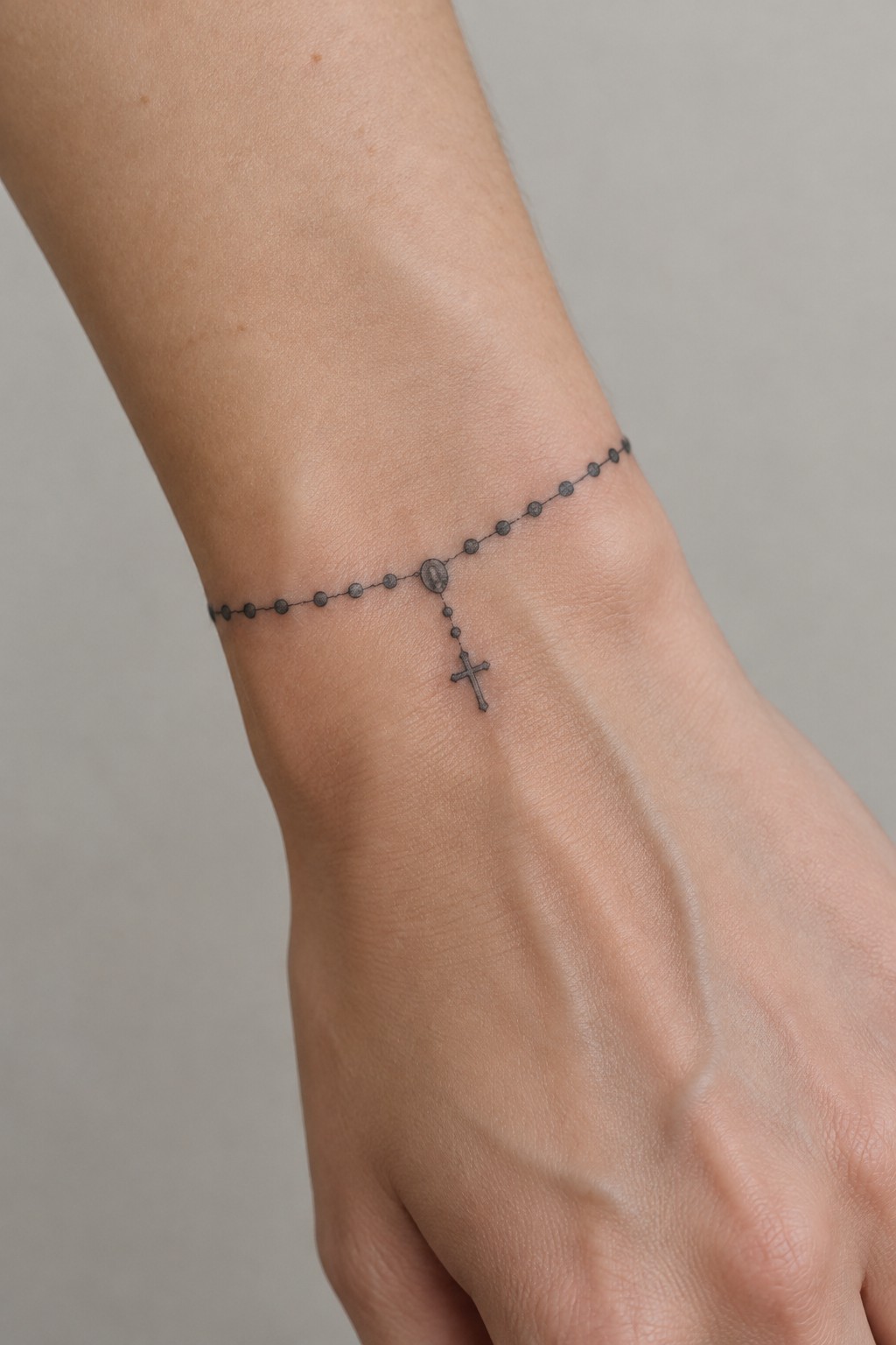 Fine-line rosary bracelet tattoo with small beads and a dangling cross around the wrist.