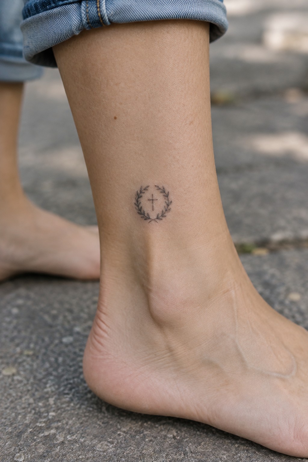 A small black fine-line cross inside a laurel wreath tattoo on the inner ankle.