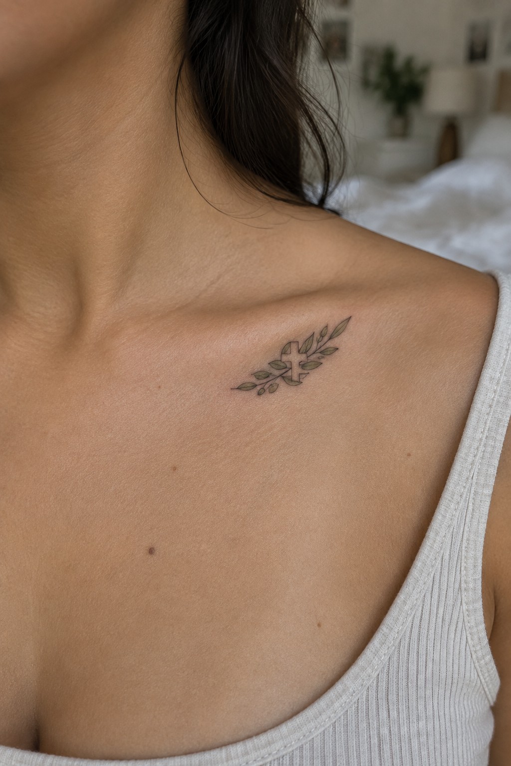 Small fine-line cross tattoo with olive branches on a woman’s upper chest near the collarbone.