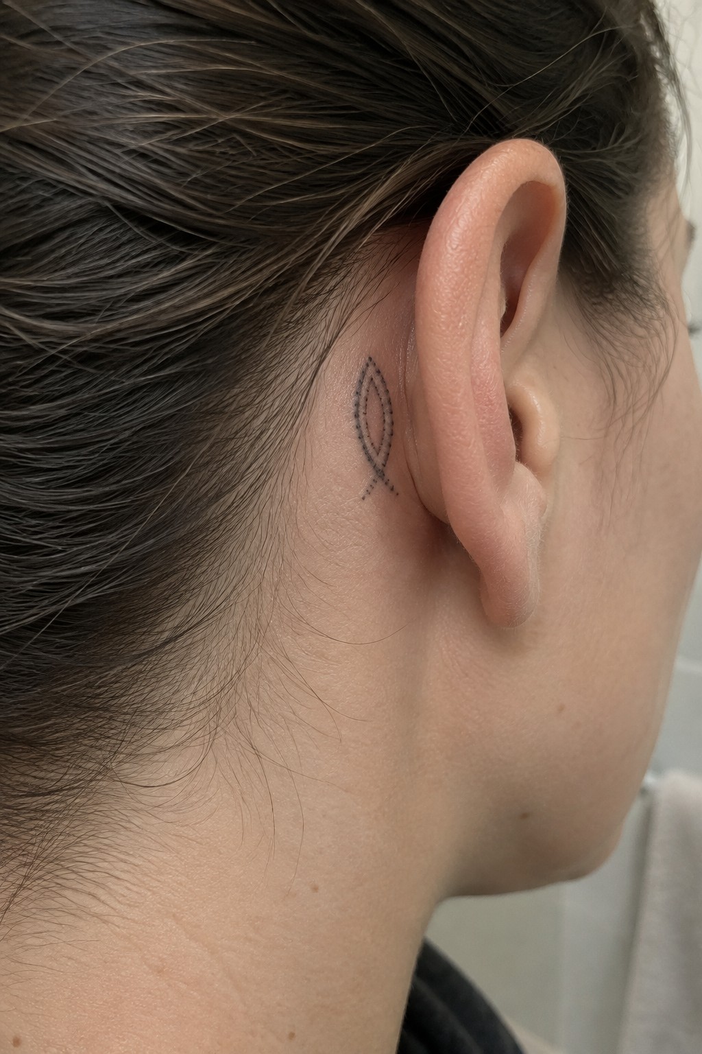Small dotted outline of a fish tattooed behind a woman’s ear.