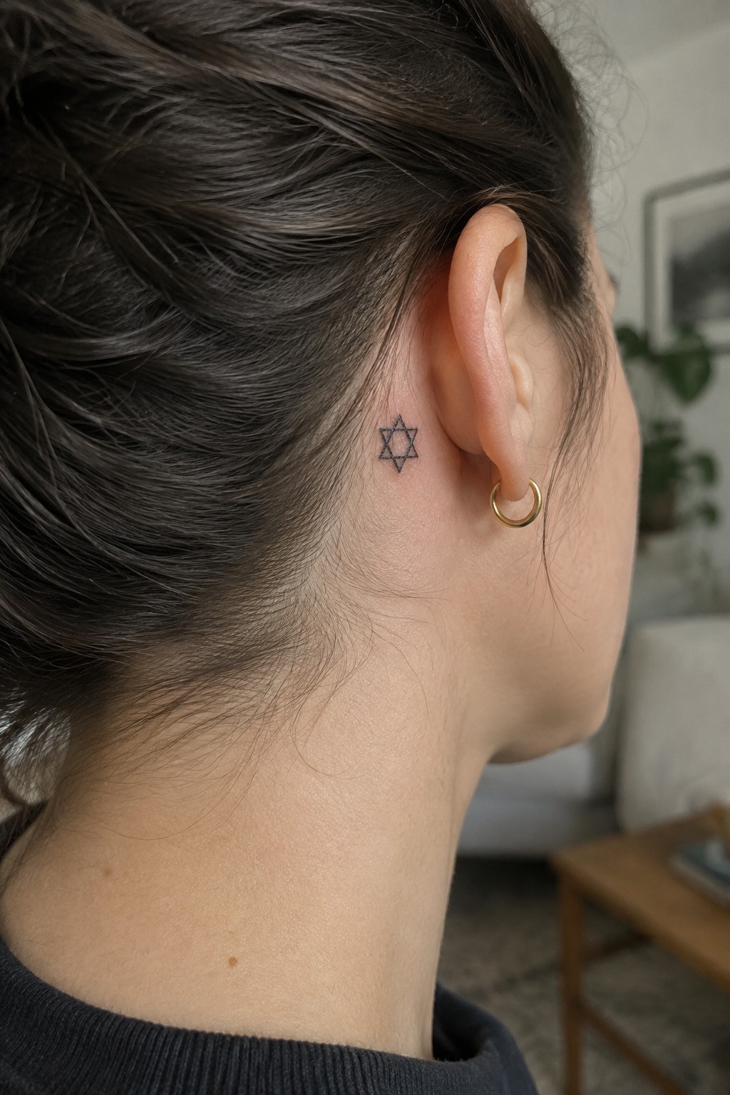 Small fine-line Star of David tattoo behind a woman’s ear, above a gold hoop earring.