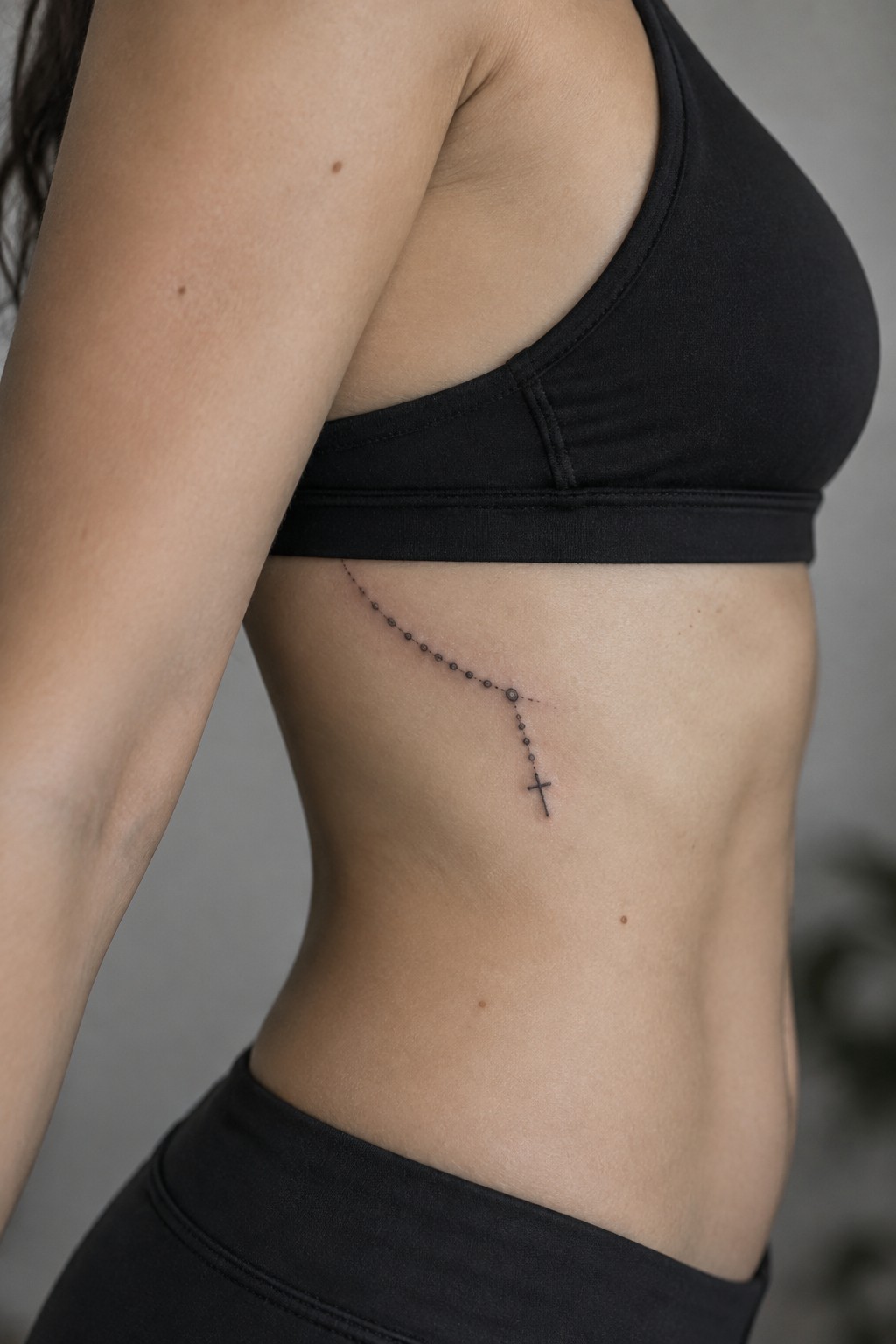 Fine-line rosary tattoo with a small cross along a woman’s side ribs beneath a black sports bra.