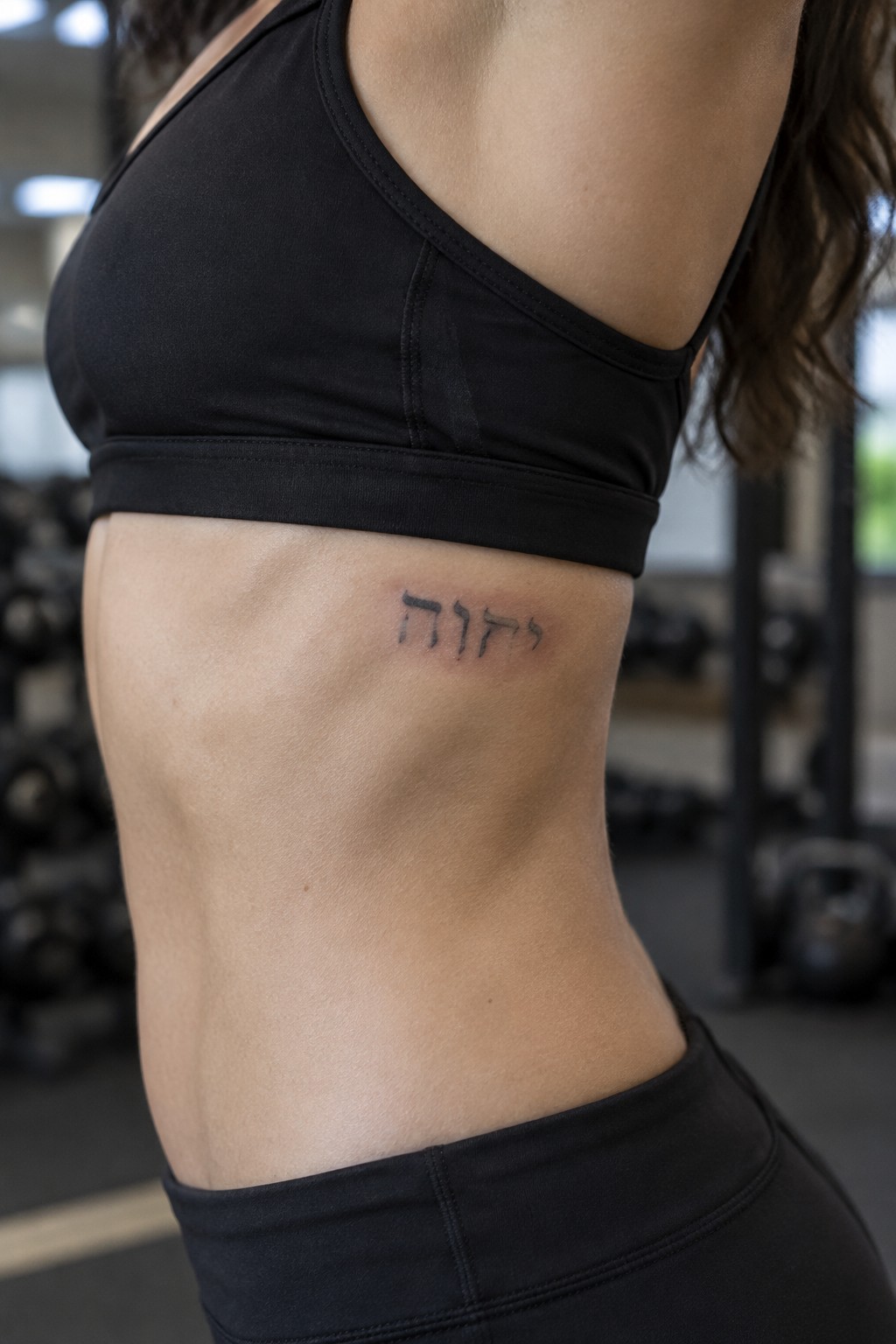 Woman's athletic torso with Hebrew tattoo below black sports bra in gym