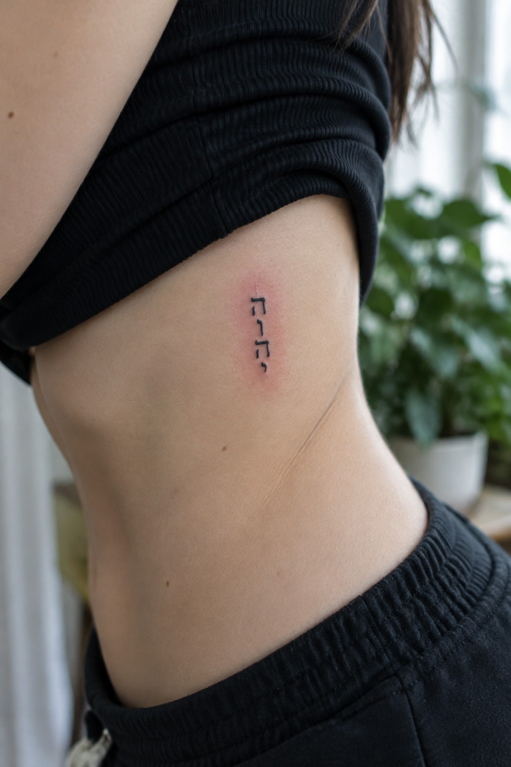 Close-up of small black tattoo on skin under lifted black ribbed top with blurred plants behind.