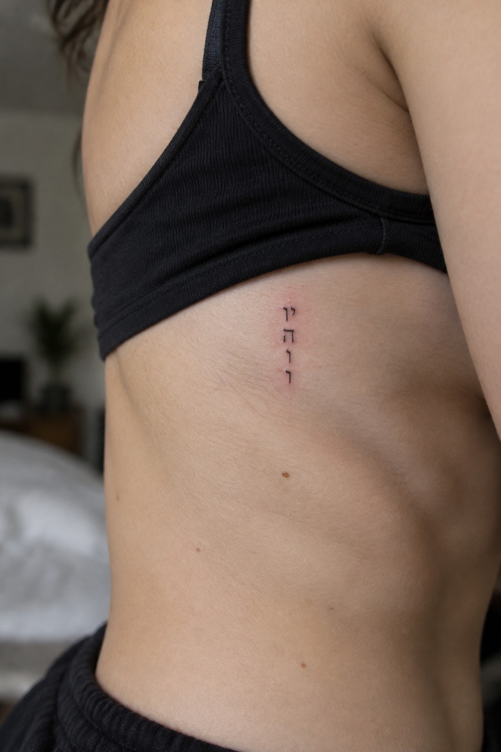 Torso closeup with vertical Hebrew tattoo on skin below black crop top.