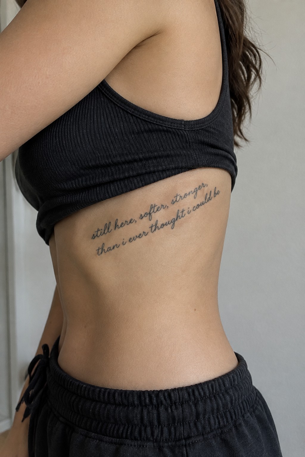 Fine-line cursive tattoo reading “still here, softer, stronger than i ever thought i could be” along the side of the ribs.