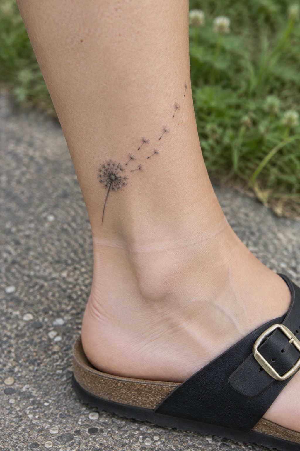 Small black dandelion tattoo with scattered seeds on the lower leg above a sandal.