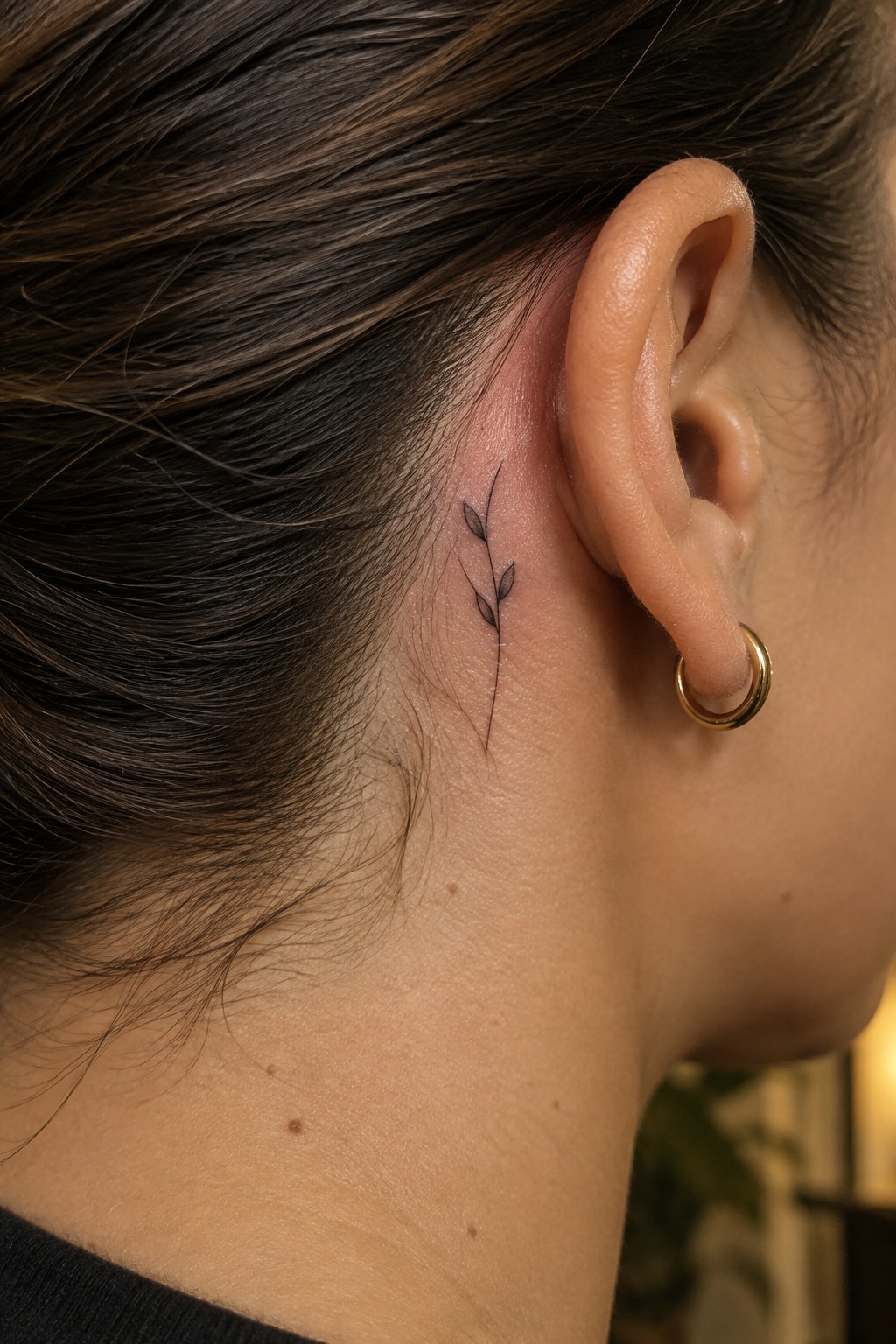 Small fine-line sprig tattoo behind a woman’s ear, with a gold hoop earring.