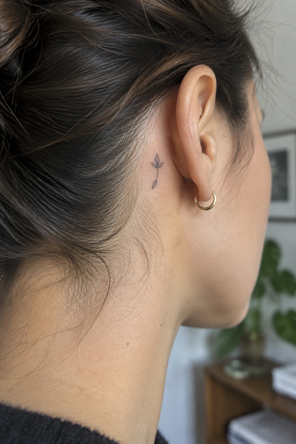 Small fine-line sprig tattoo behind a woman’s ear, above a gold hoop earring.