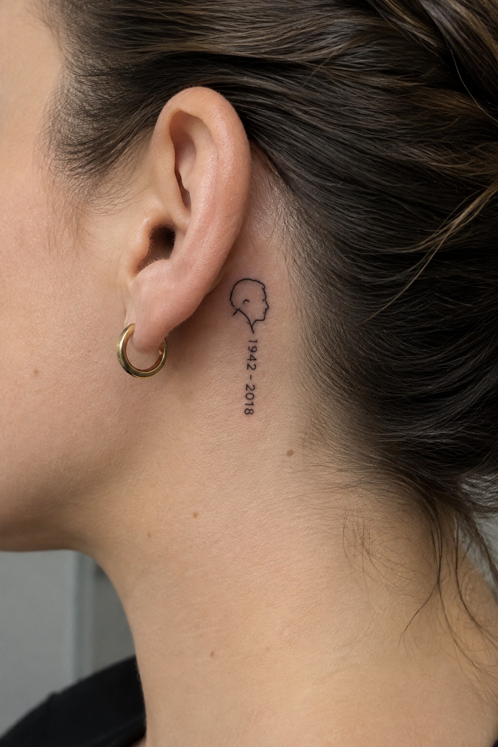Small black outline profile portrait tattoo with the dates 1942–2018 behind a woman’s ear.