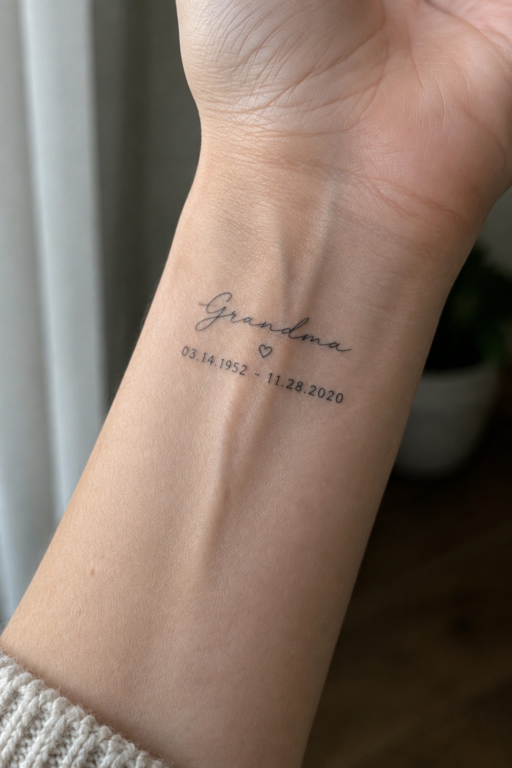 Small black script tattoo reading “Grandma” with a heart and the dates 03.14.1952 and 11.28.2020 on the inner wrist.