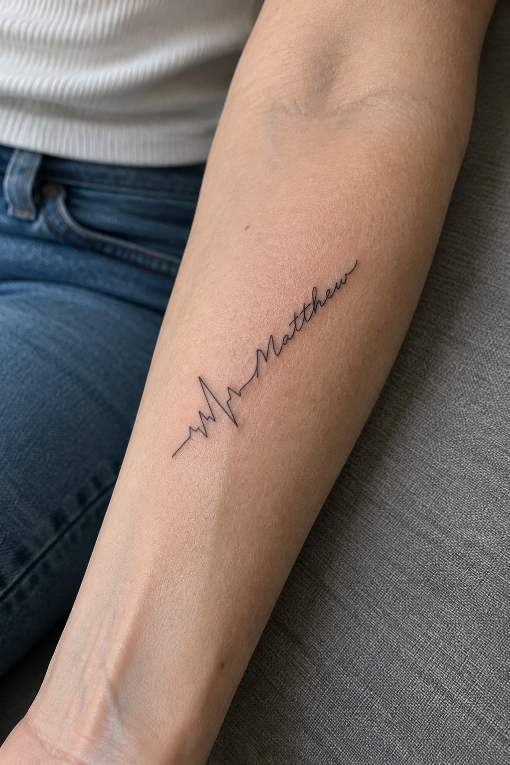 Fine-line ECG tattoo leading into the name Matthew on the inner forearm.