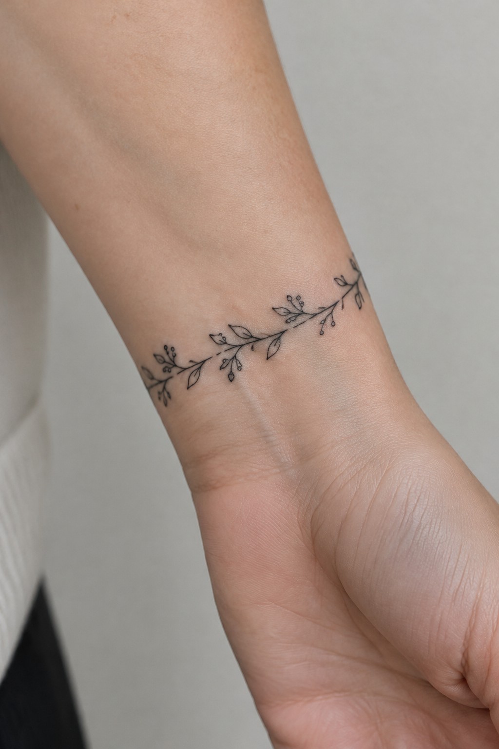 Fine line botanical vine tattoo with leaves and buds wrapping around the wrist