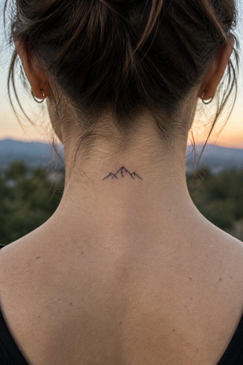 Woman's neck with small mountain tattoo, brown hair in bun, sunset backdrop
