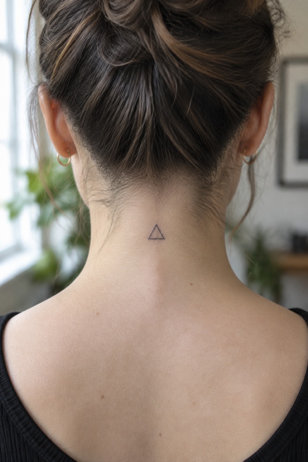 Close-up of a woman's neck with a tiny black triangle outline tattoo centered on the nape below a hair bun.