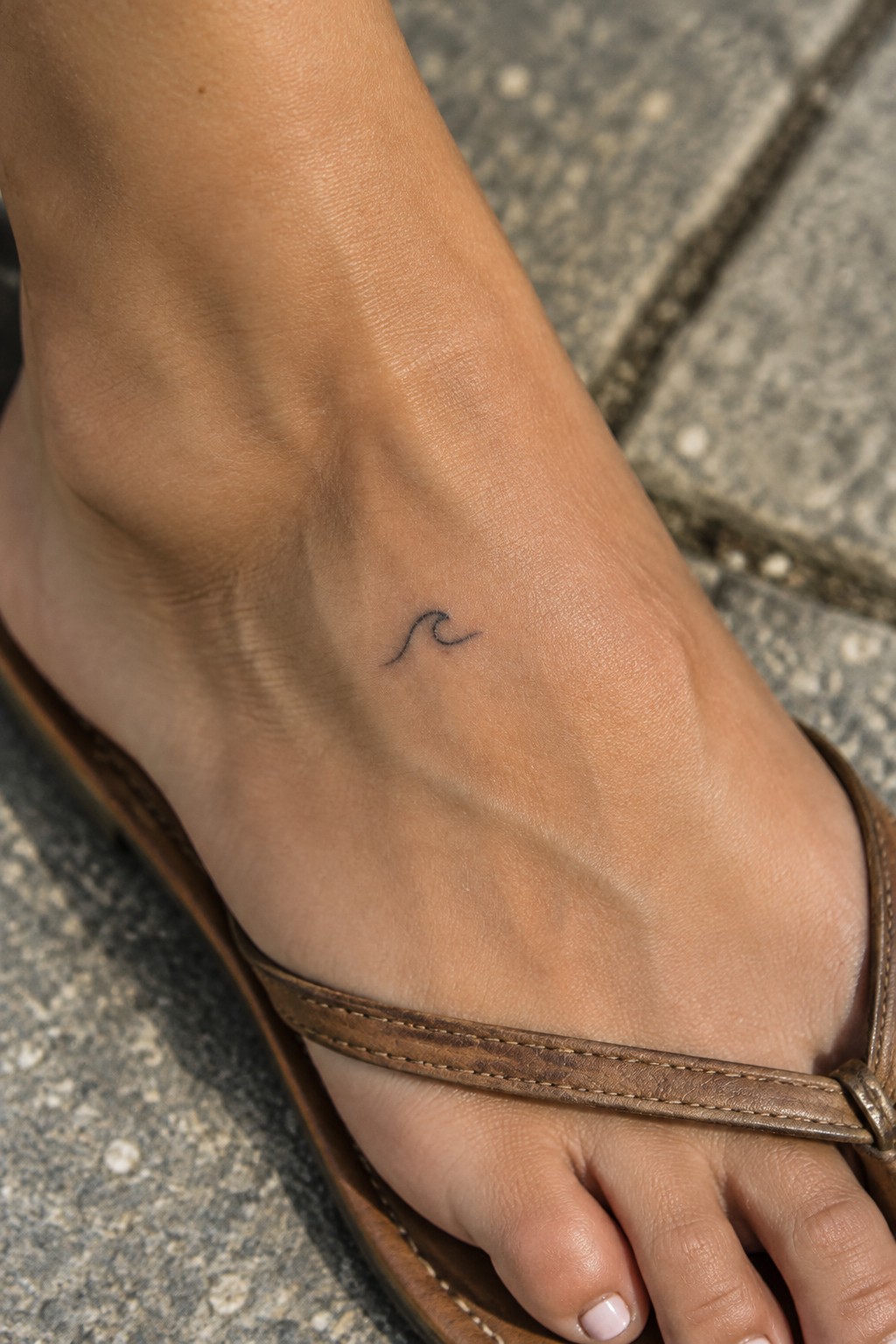 Foot with small wave tattoo in brown leather flip-flop on stone pavement.
