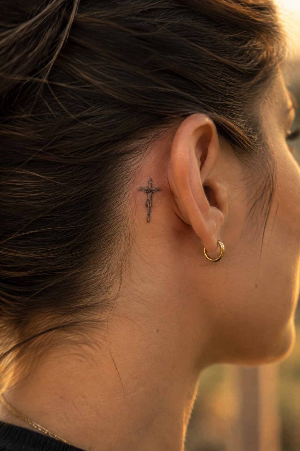 Crucifix tattoo behind woman's ear with gold hoop earring and thin necklace.