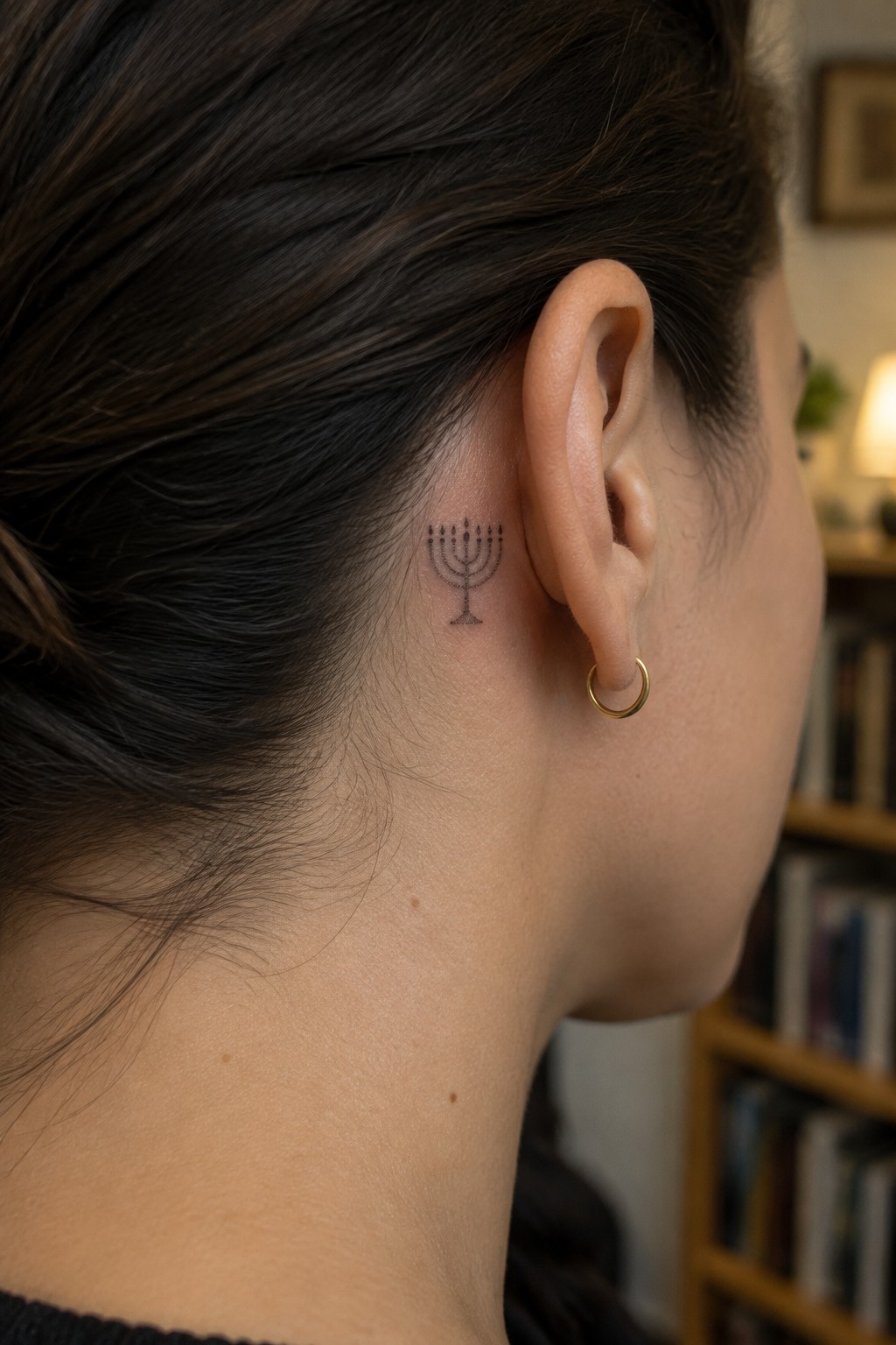 Close-up of neck with menorah tattoo behind ear and small gold hoop earring.