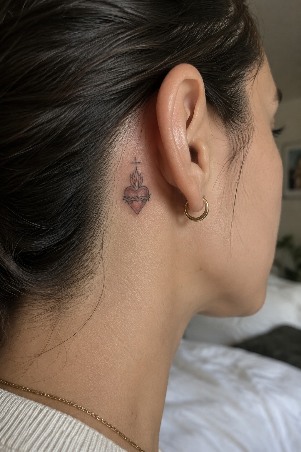 Sacred heart tattoo behind ear with gold hoop earring, necklace, and white top