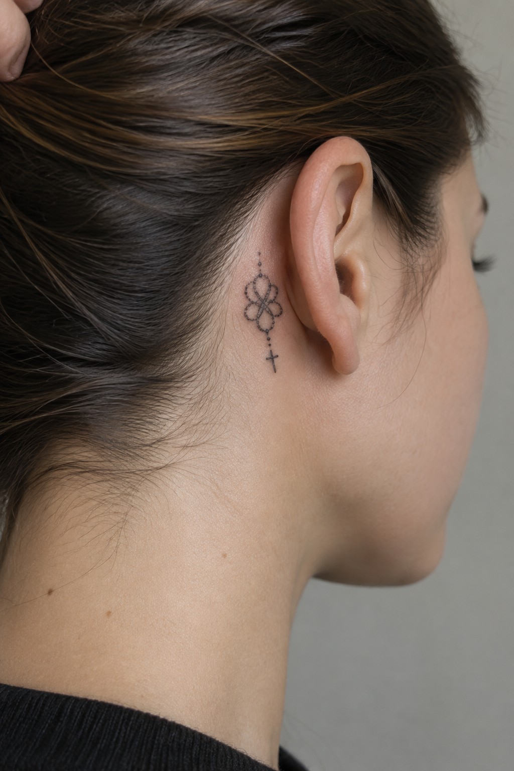Small black dotwork tattoo of a cross hanging from a looped floral motif behind a woman's ear