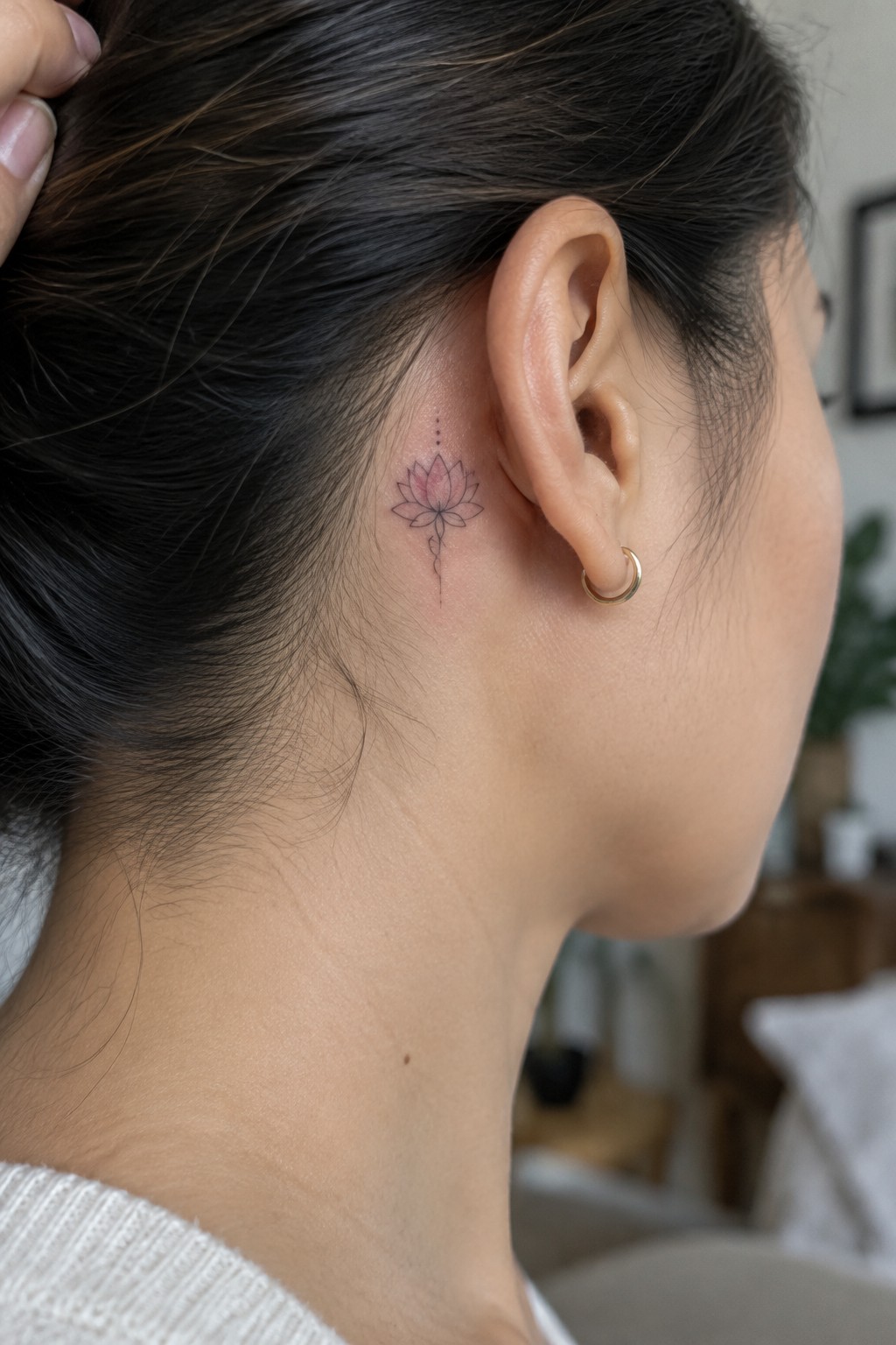 Pink lotus tattoo behind ear on woman with dark bun and gold hoop earring