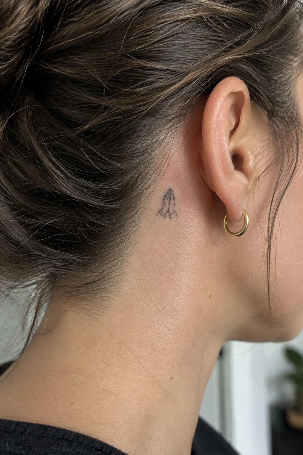 Small fine line tattoo of praying hands positioned behind a woman's ear on the side of her head.