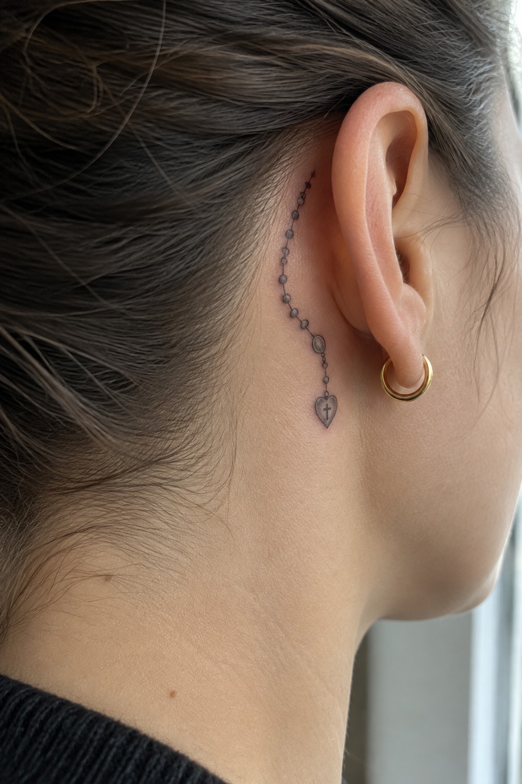Close up of a small rosary tattoo behind a woman's ear, made of fine dotted beads that curve downward and end in a heart pendant with a cross inside it.