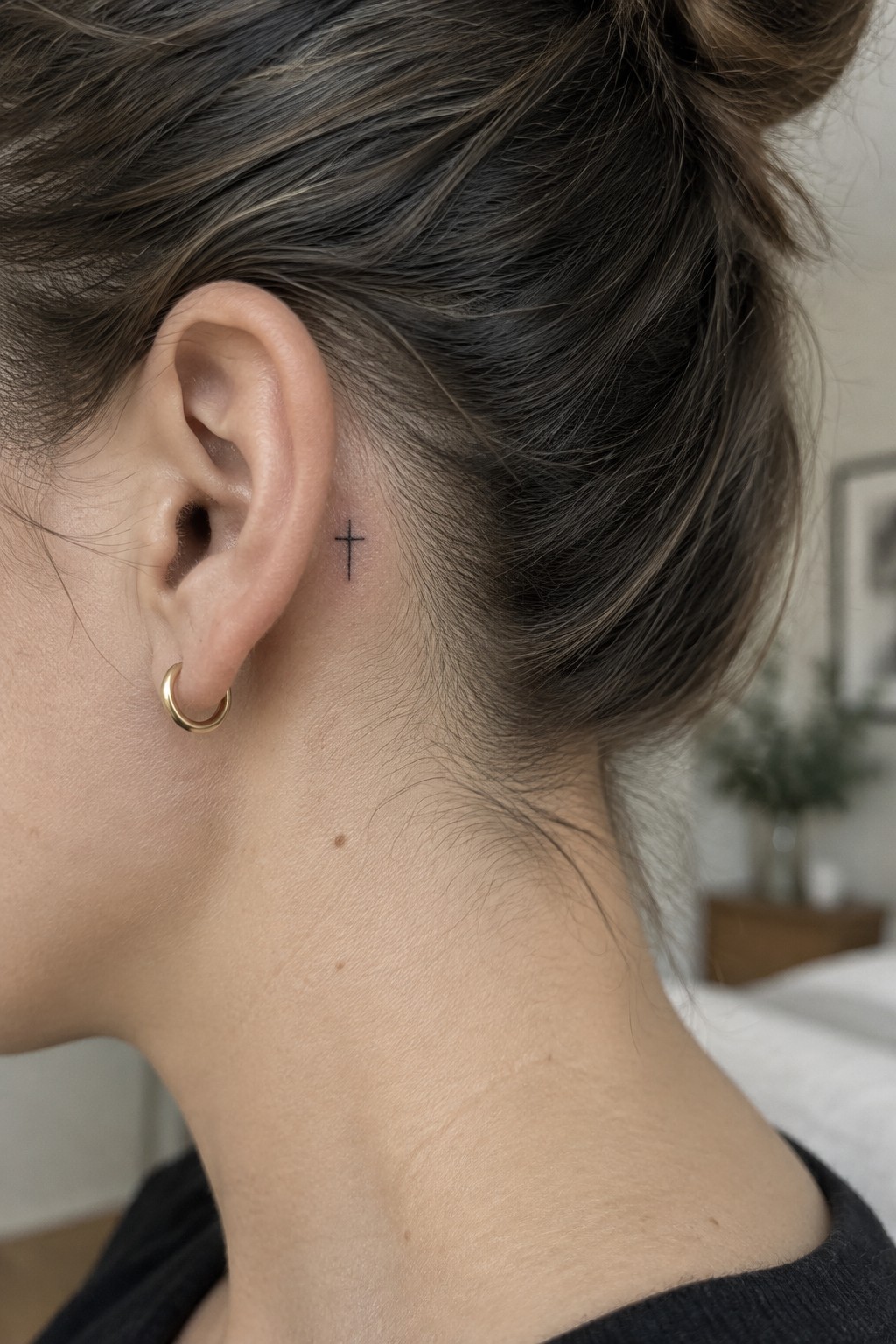 Small black cross tattoo placed behind the ear on a woman with hair in a bun and a gold hoop earring.