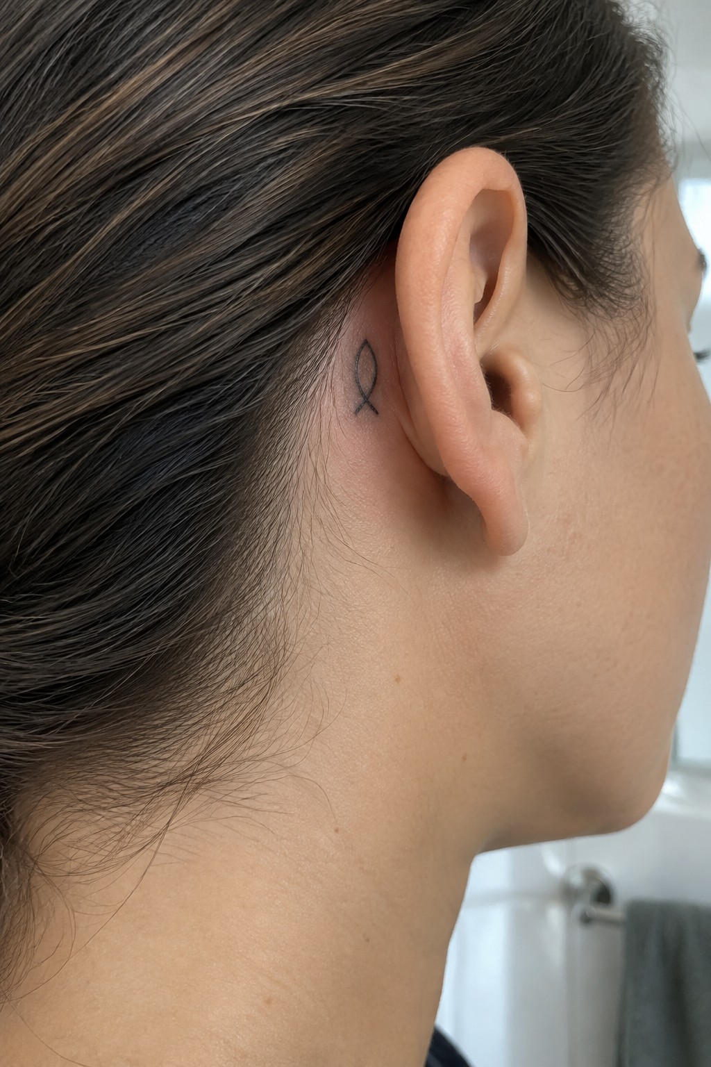 Close-up side view of ear with small black fish tattoo behind it and dark hair.