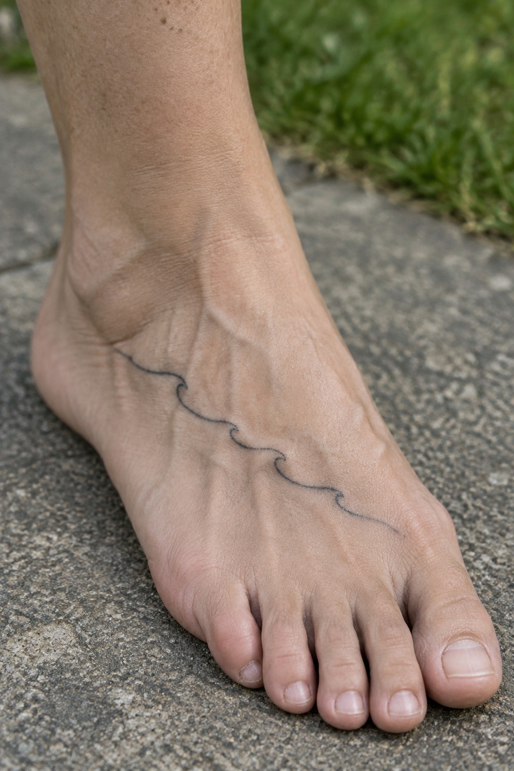 Bare foot with wavy black tattoo on gray stone near green grass