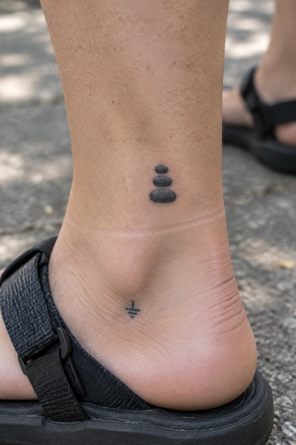 Black stacked stones tattoo on ankle above small geometric symbol, foot in black sandal.