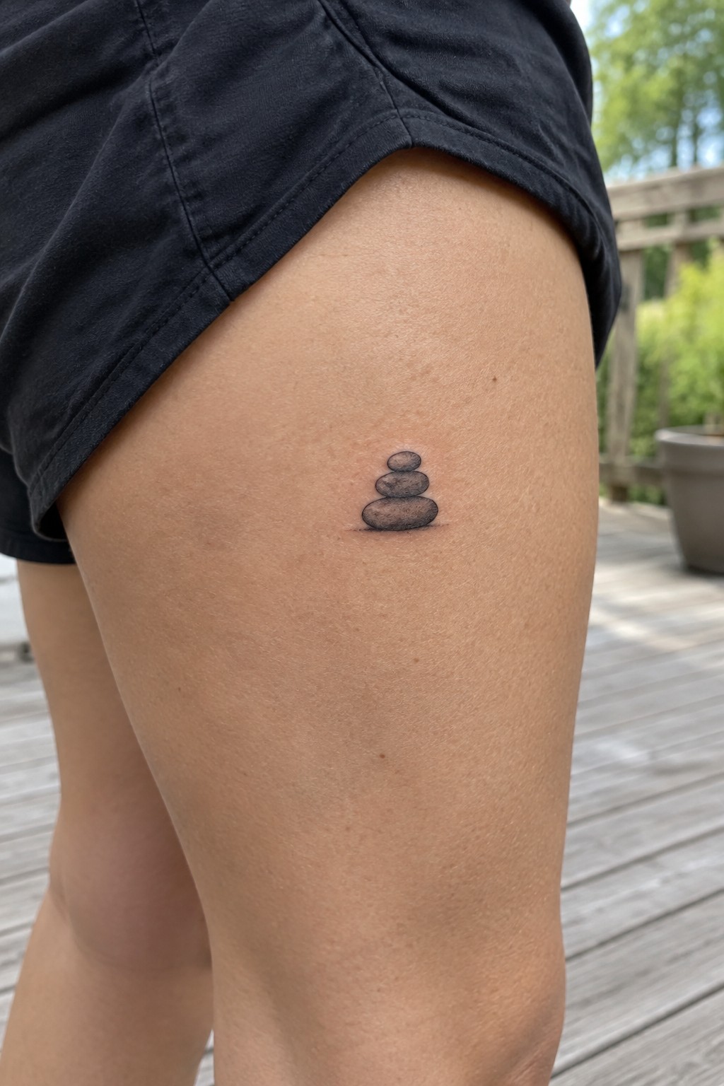 Small black and gray tattoo of three stacked stones on the upper thigh, shown on light skin with natural shading.